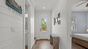 Un pasillo con paredes de madera blanca, un banco de madera y una puerta que da a un dormitorio con cama y ventana.