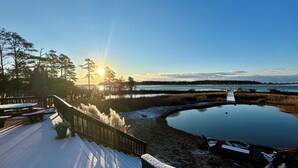 Ein mit Schnee bedeckter Deck mit Picknicktischen, einem Holzgeländer und Blick auf einen ruhigen See, dessen hölzerner Steg sich ins Wasser erstreckt.