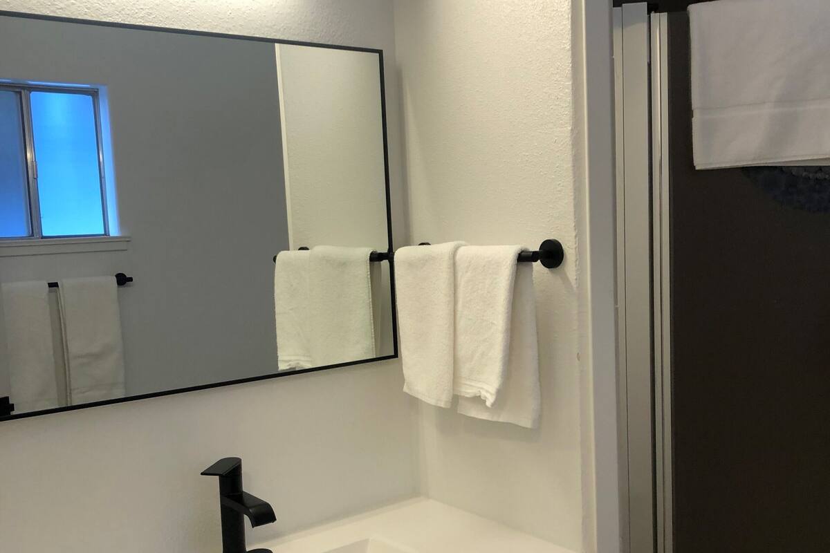 Ein Badezimmer mit einem weißen Waschbecken, einem schwarzen Wasserhahn und einem Spiegel darüber. Zwei Handtücher hängen an einem Handtuchhalter.