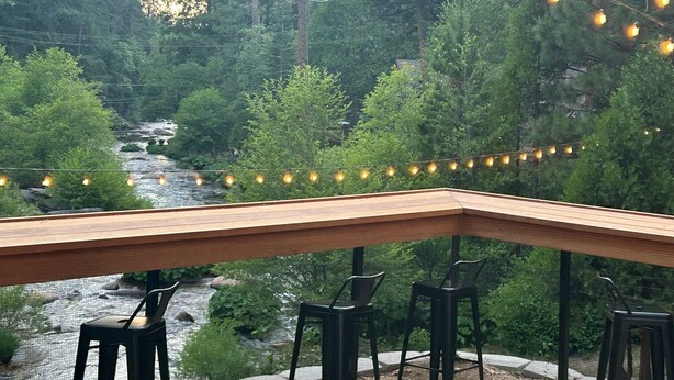 A wooden deck with chairs overlooking a river, surrounded by trees and string lights. - Sierra Inn on the River (Kyburz)