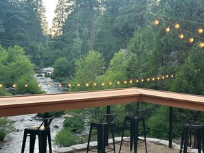 A wooden deck with chairs overlooking a river, surrounded by trees and string lights. - Sierra Inn on the River (Kyburz)