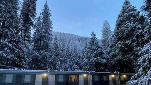 A multi-story building with a row of rooms, surrounded by snow-covered trees and a forested hill in the background. - Sierra Inn on the River (Kyburz)