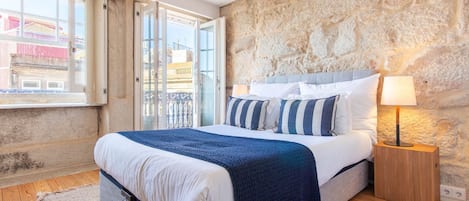A bedroom with a stone wall, a bed with striped pillows, a nightstand with a lamp, and a view of buildings through a window.