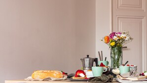 Una mesa con desayuno que incluye pan, fruta, una cafetera y un florero.