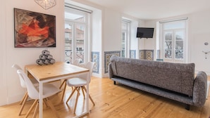 A modern living room with a wooden dining table, white chairs, a grey sofa, and a flat-screen TV mounted on the wall. - Lusitano's 2BDR Apartment in Alfama by LovelyStay (Lisboa)