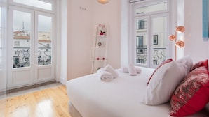 A hotel room with a bed, a small shelf with decorative items, and a view of the city through the windows. - Lusitano's 2BDR Apartment in Alfama by LovelyStay (Lisboa)