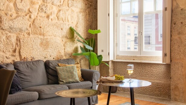 Una sala de estar acogedora con pared de piedra, un sofá gris, una planta verde y una pequeña mesa redonda con una copa de vino.