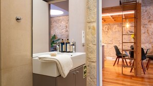 Un baño con lavamanos, espejo y artículos de aseo. Una puerta de madera da a una zona de comedor con sillas y una mesa.