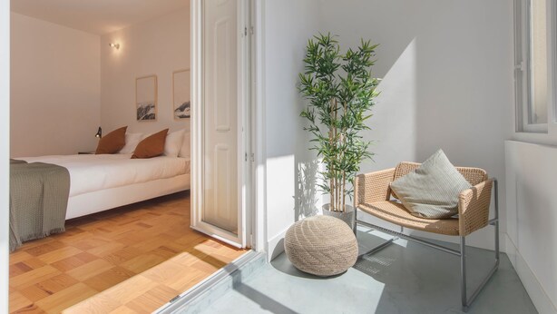 Ein Schlafzimmer mit einem Bett, einem Sessel, einer Topfpflanze und einer Tür zu einem anderen Raum.