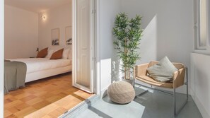 A bedroom with a bed, a chair, a potted plant, and a door leading to another room.