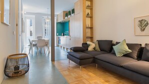 Ein modernes Wohnzimmer mit einem dunkelgrauen Sofa, einem Essbereich mit weißem Tisch und Stühlen sowie einer Küche mit türkisfarbener Spritzschutzleiste.