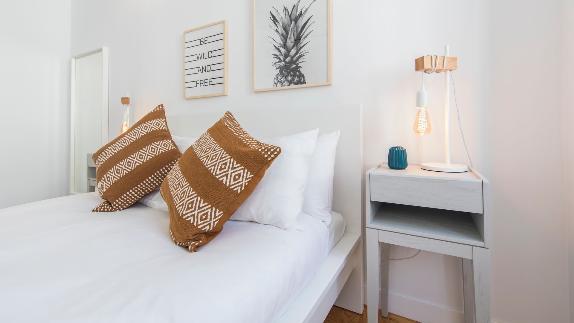 A neatly made bed with patterned pillows, a bedside table with a lamp, and wall art including a pineapple and a motivational quote.