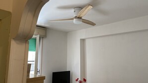 Una habitación con ventilador de techo, un televisor de pantalla plana sobre un soporte de madera, un florero con flores rojas y una ventana con cortinas.