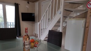 Une pièce avec une table en verre sur laquelle sont disposés une bouteille et des verres, un panier de fruits et une chaise. Un téléviseur est fixé au mur, et un escalier est visible au fond.