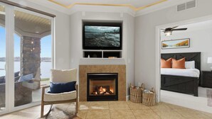 Un salon moderne avec une cheminée, un téléviseur fixé au-dessus, un lit avec une tête de lit sombre et une vue sur l’extérieur grâce à une porte vitrée.