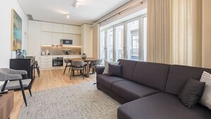 A modern living room with a grey sofa, a dining table with chairs, a kitchen area with a microwave and cabinets, and large windows with curtains. - Hidden Oasis 2BDR Apartment by LovelyStay (Lisboa)