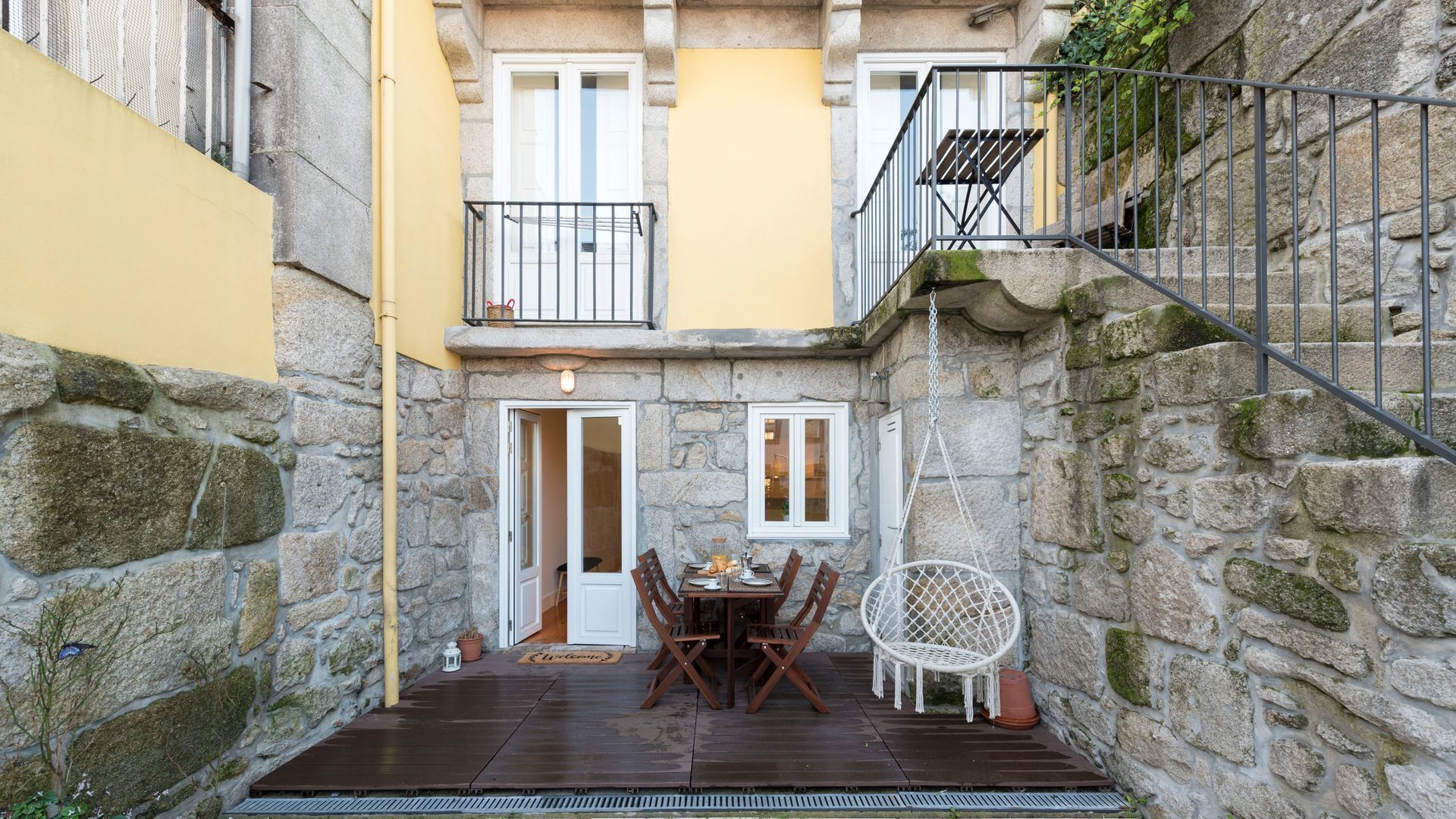 A courtyard with a wooden table and chairs, a hanging chair, and a stone wall.
