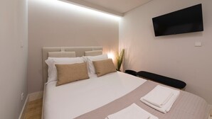 Een net opgemaakt bed met wit en beige beddengoed, een flatscreen televisie aan de muur bevestigd, en een nachtkastje met een lamp.