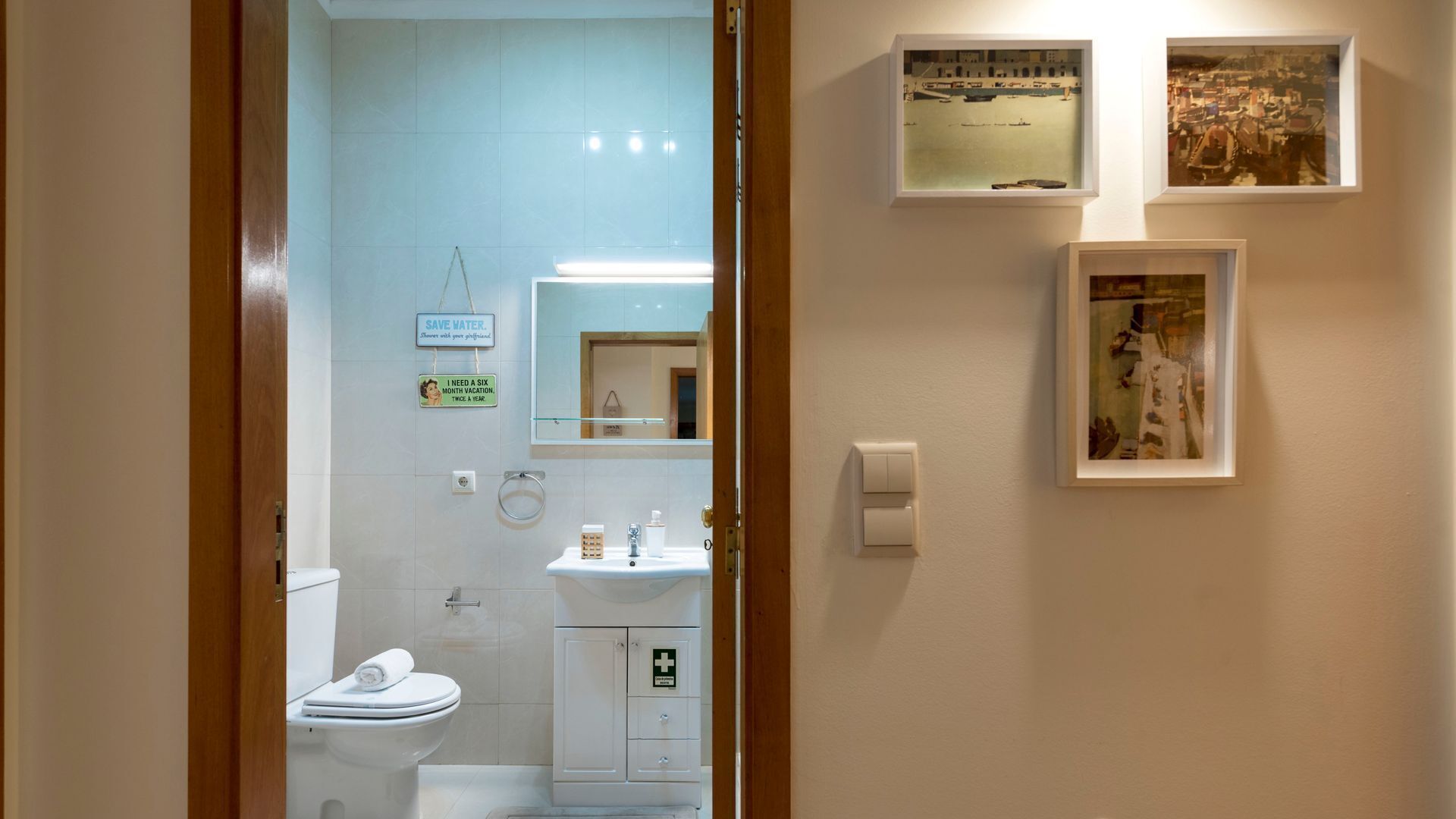 A bathroom with a toilet, sink, and a mirror. There are two framed pictures on the wall.
