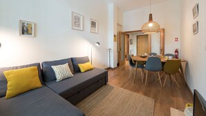 Uma sala de estar moderna com um sofá cinza, almofadas amarelas e estampadas, uma área de jantar com mesa e cadeiras de madeira, e um lustre.