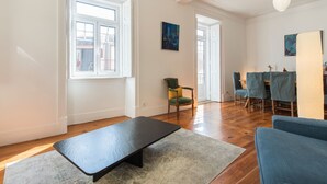 Een ruime woonkamer met houten vloeren, een zwarte bank, een groene fauteuil, en een eethoek met blauwe stoelen.