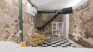 Una habitación con paredes de piedra, suelo a cuadros, sillas de mimbre, una cama y una escalera.