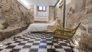 Una habitación con piso a cuadros, una cama con cubierta blanca, sillas de mimbre y un ventanal pequeño.