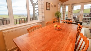 A wooden dining table with chairs, a fruit bowl, and a view of the countryside through large windows. - Lodge 23 (Torpoint)