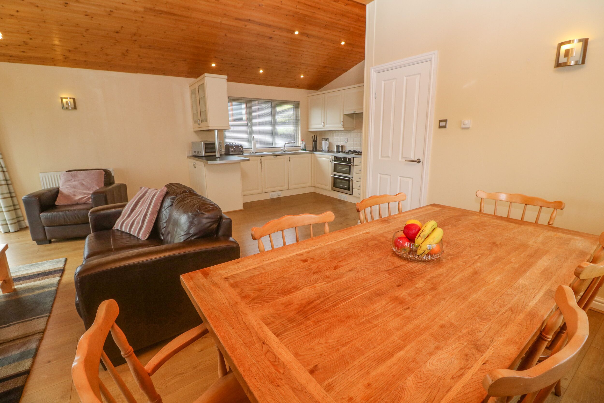 A cozy living room with a brown leather sofa, a wooden dining table with chairs, and a fruit bowl on the table.