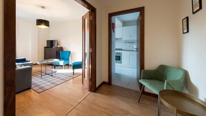 Ein modernes Wohnzimmer mit einer Couch, Sesseln, einem Couchtisch und einem karierten Teppich. Durch eine offene Tür ist eine Küche zu sehen.