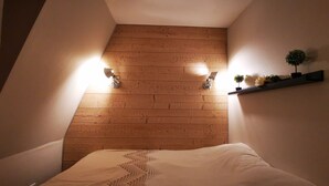 A bedroom with a sloped ceiling, wooden paneling, a bed with a white duvet, two wall-mounted lamps, and a shelf with a plant and flowers. - NEW - ULTRA CENTER & PANORAMIC VIEW (Huez)
