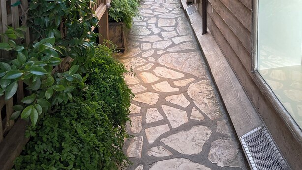 Un estrecho callejón empedrado de piedra con una pérgola de madera, bordeado por exuberante vegetación y vallas de madera.