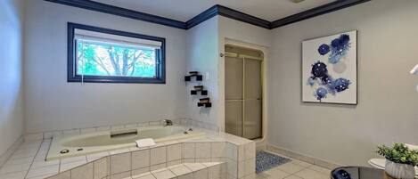 A bathroom with a large bathtub, a walk-in shower, a window, and a framed abstract painting.