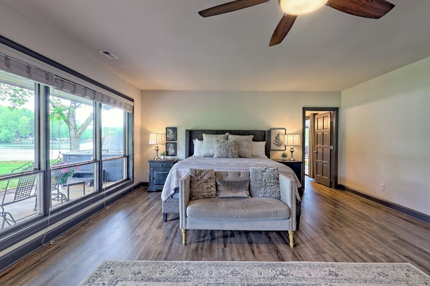A bedroom with a large window, a ceiling fan, a bed with multiple pillows, a nightstand, and a chair.