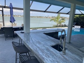 A covered outdoor bar area with a pool, bar stools, and a view of the water. - Stunning Waterfront Retreat with Private Pool and beautiful entertaining space. (Naples)