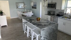 Una cocina moderna con armarios blancos, una isla de granito, taburetes de bar blancos y una zona de comedor con una mesa y sillas blancas.