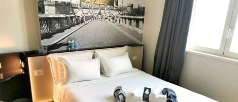 Una habitación de hotel con cama, cabecera, ventana con cortinas, un cuadro en la pared de un edificio histórico y una mesita de noche con lámpara.