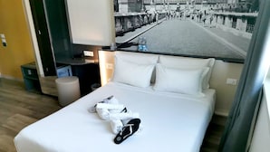 A hotel room with a bed, pillows, a TV, and a view of a street with people. - B&B Hotel Roma Fiumicino Aeroporto Fiera 2 (Fiumicino)