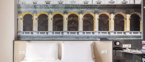 A hotel room with a bed, a desk with a lamp, and a mural of an architectural facade.
