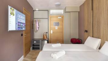 A hotel room with two beds, a TV displaying a hotel app, a wooden wardrobe, a desk with a handbag, and a red suitcase.