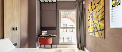 A hotel room with a bed, a desk, a chair, a window, and a wall with a painting.