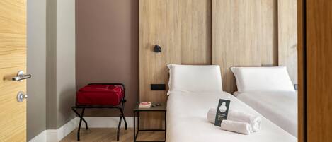 A hotel room with a wooden wardrobe, a small bedside table with a red bag, and neatly made beds.