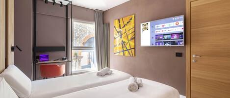 A hotel room with two beds, a desk with a laptop, a TV displaying streaming services, and a large window with curtains.