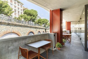 Een balkon met twee tafels en stoelen, een stenen muur met bogen, en uitzicht op gebouwen en bomen.