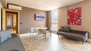 A modern living room with a sofa, a dining table with chairs, a TV mounted on the wall, and a large abstract painting.