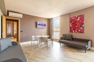 Een moderne woonkamer met een bank, een eettafel met stoelen, een aan de muur bevestigde televisie en een groot abstract schilderij.
