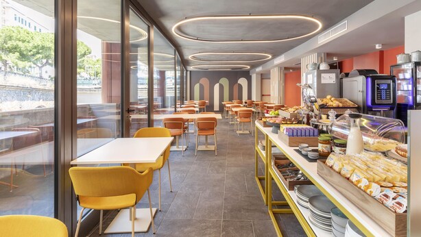 A modern cafeteria with tables and chairs, a buffet area with food items, and a view of the outdoors through large windows.