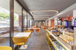 Een moderne cafeteria met tafels en stoelen, een buffetgedeelte met etenswaren en uitzicht op het buitenleven door grote ramen.