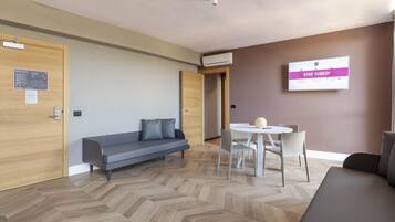 Una habitación de hotel moderna con una puerta de madera, un sofá gris, una mesa de comedor redonda con sillas y un televisor montado en la pared que muestra “STAY TUNED!”.