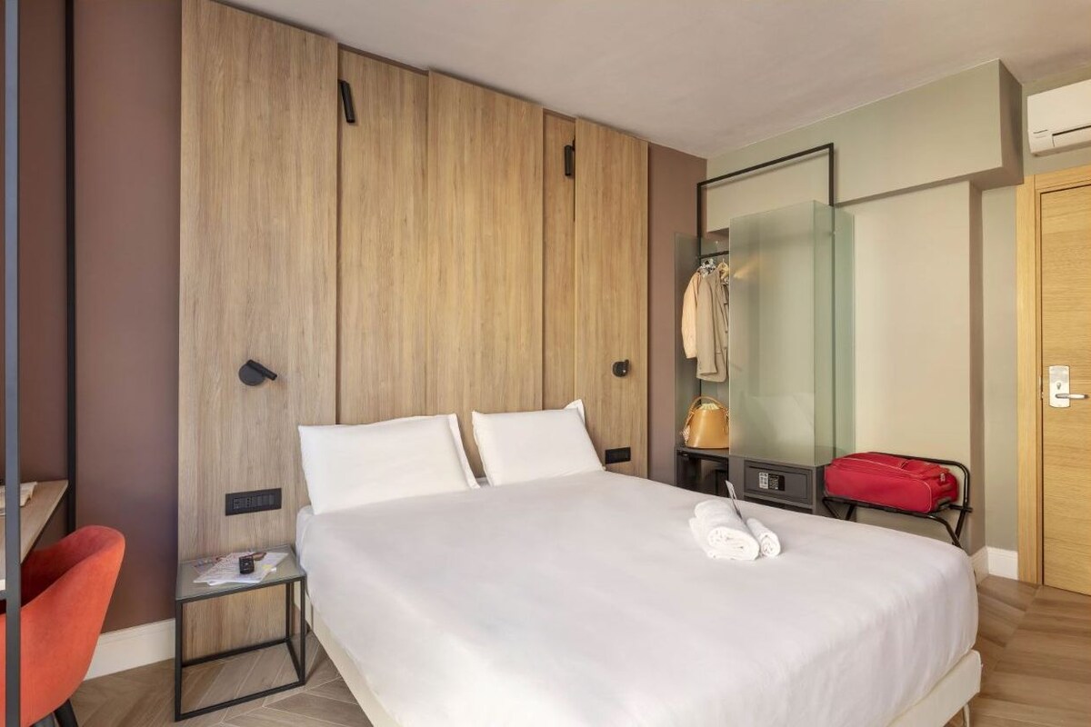 Habitación de hotel con una cama grande, un armario de madera, un espejo, una silla roja y una maleta roja.